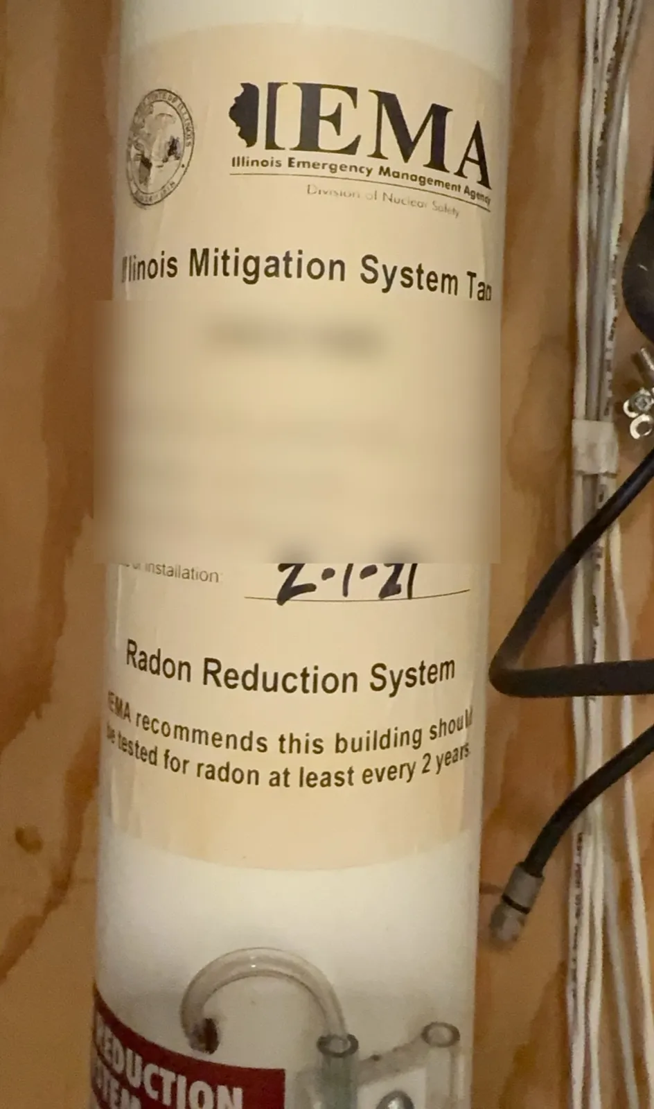 Illinois Mitigation System Tag on a white radon vent pipe, reading Radon Reduction System with a handwritten installation date; the tag number and installer details are blurred