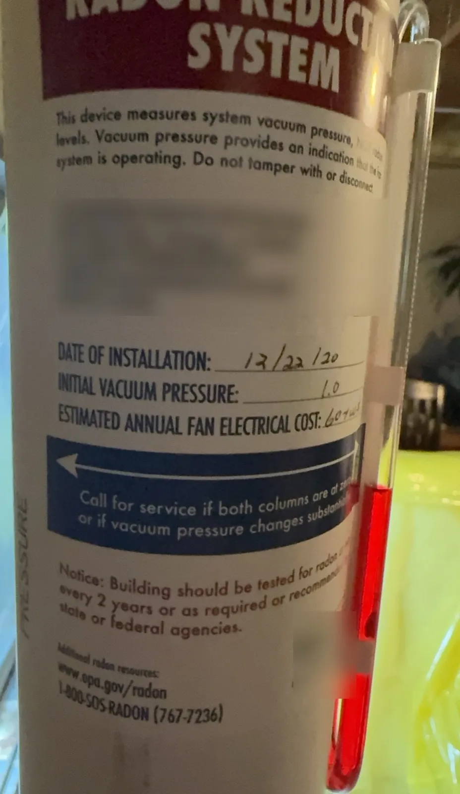 U-tube manometer with red fluid on a radon vent pipe, beside a label recording the installation date and the initial vacuum pressure; the installer's details are blurred